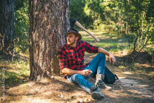 Handsome young man with axe near forest. Lumberjack sitting in the forest.