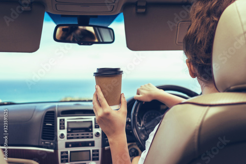 Fototapeta Naklejka Na Ścianę i Meble -  Woman driver drinking coffee paper cup inside car during driving. Girl relaxing in auto trip traveling along ocean tropical beach in background. Traveler concept. Back view