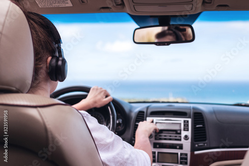 Woman driver in the headphones driving a car. Girl relaxing in auto trip traveling along ocean tropical beach in background. Traveler concept. Back view © flowertiare