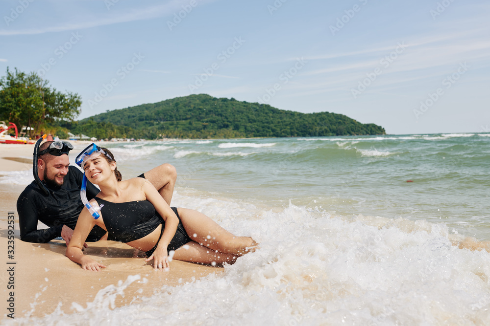 Happy young couple lying on beach and enjoying warm sea waves after snorkeling together