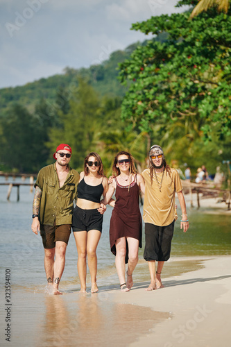 Two smiling couples in sunglasses hugging when walking on tropical beach together
