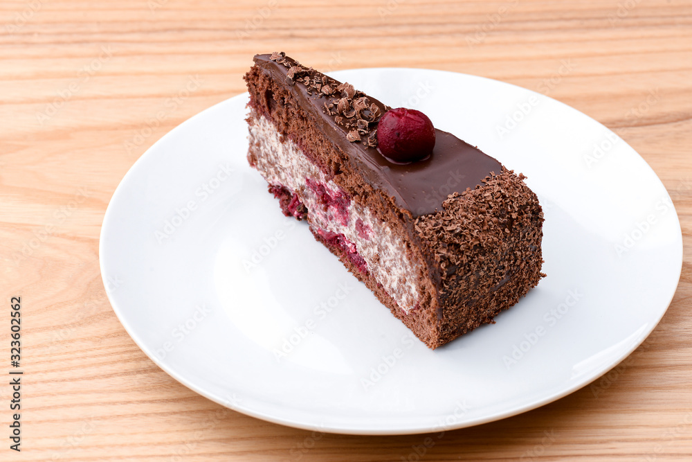 One piece of chocolate cake with vanilla cream and cherry topping served on white plate over light wooden background.
