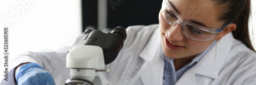 Portrait of research technician putting some substance under microscope in laboratory office. Smiling woman in protective uniform. Science and investigation concept