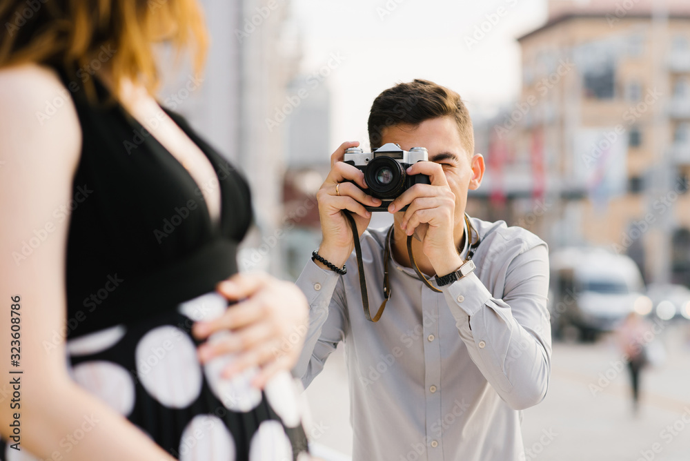 A beautiful stylish couple of future parents are walking around the city. The future father photographs the future mother A beautiful stylish couple of future parents are walking around the city. The future father photographs the future mother