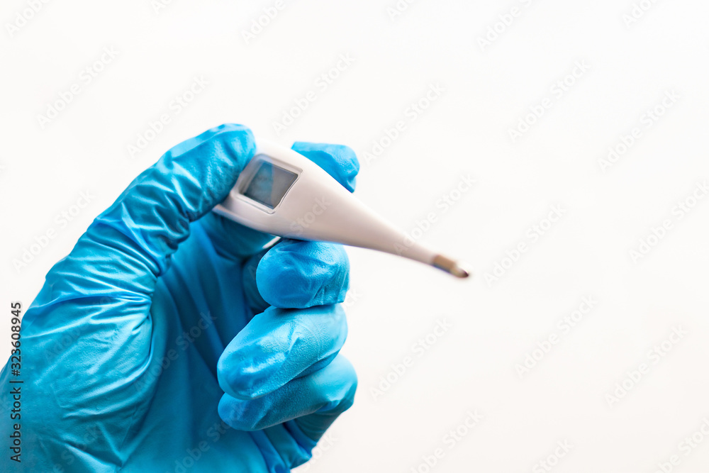 A doctors hand in a glove holds a electronic thermometer close up on a light background with copy space. High temperature, symptoms of a virus or illness. The diagnosis of health in the clinic. A doctors hand in a glove holds a electronic thermometer close up on a light background with copy space. High temperature, symptoms of a virus or illness. The diagnosis of health in the clinic.