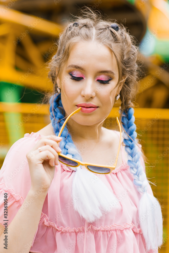 A beautiful girl with blue pigtails and bright pink and lilac professional makeup holds a pair of yellow-rimmed sunglasses near her lips A beautiful girl with blue pigtails and bright pink and lilac professional makeup holds a pair of yellow-rimmed sunglasses near her lips