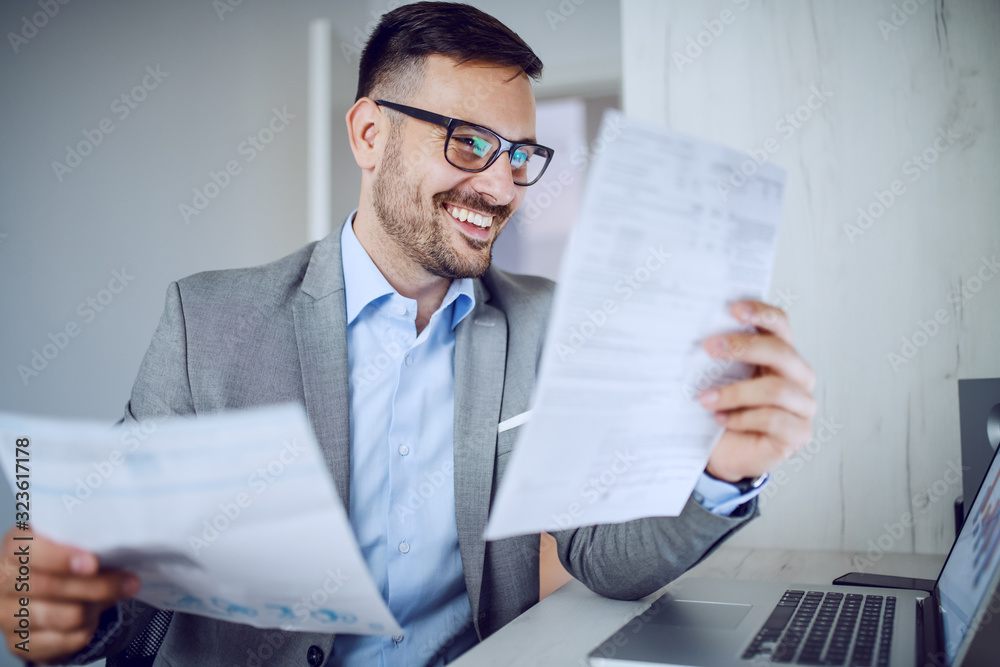 Hardworking sophisticated caucasian businessman in suit and with eyeglasses looking at documents while sitting in his office. On desk is laptop.