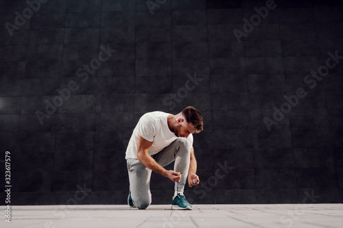 Handsome Caucasian bearded blonde muscular man in tracksuit kneeling and tying shoelace. In background is gray wall.