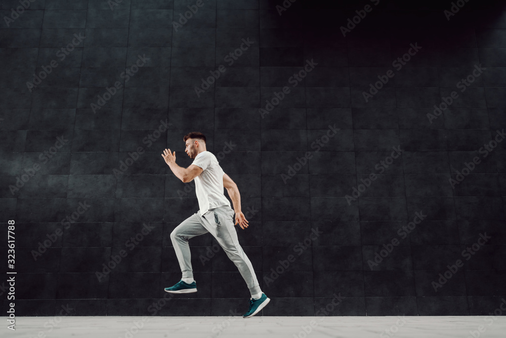 Attractive caucasian muscular fit blond bearded man in tracksuit and t-shirt running outdoors next to dark background.