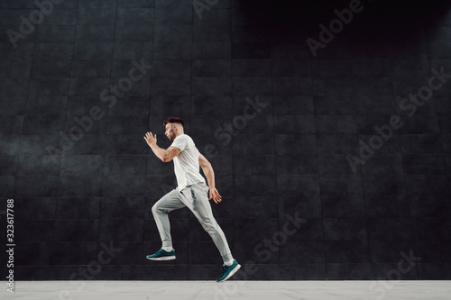 Attractive caucasian muscular fit blond bearded man in tracksuit and t-shirt running outdoors next to dark background.