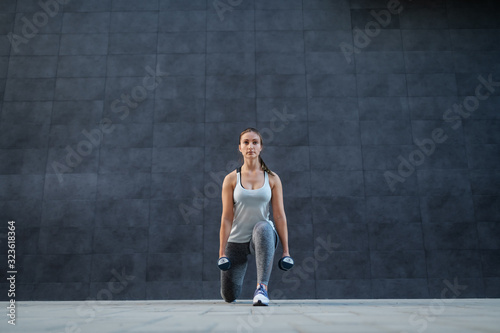 Beautiful caucasian brunette in sportswear doing lunges with dumbbells in hands and looking at camera. In background is dark wall.