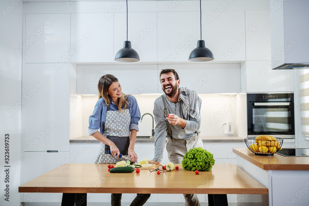 © Dusan Petkovic - Young charming smiling caucasian woman in apron standing in kitchen and cutting cucumber while talking with her boyfriend. Man holding cherry tomato and talking about healthy lifestyle.