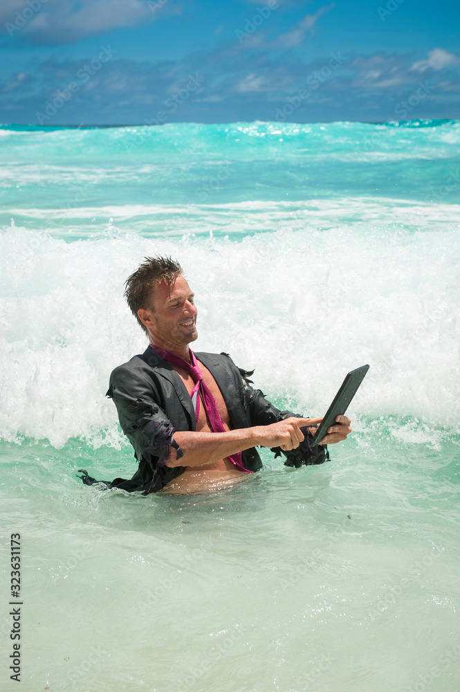Cheerful castaway businessman using a tablet computer in tropical waves