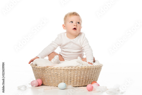 Kid with open mouth sitting on blanket in basket with Easter eggs around on white background