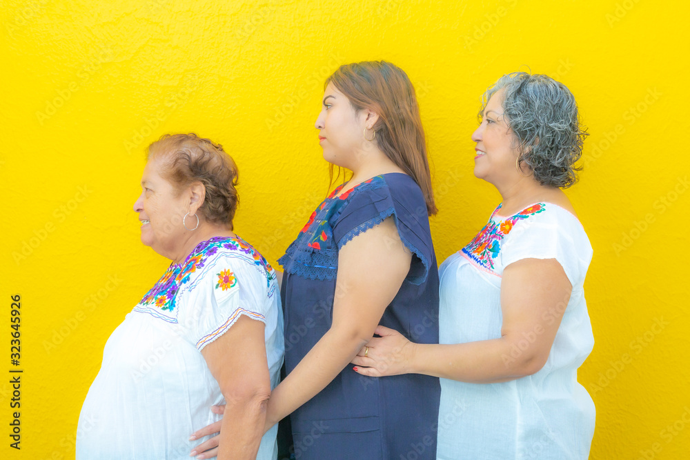 Side view profiles of three generations of Mexican women smiling with floral print blouses in a row, grandmother, granddaughter and daughter on a yellow background Side view profiles of three generations of Mexican women smiling with floral print blouses in a row, grandmother, granddaughter and daughter on a yellow background