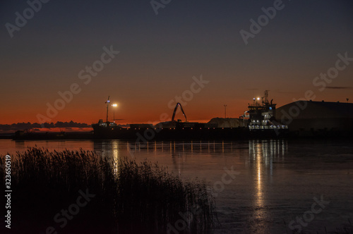 Silhouette of working crane in sea port near Parnu bay in cold winter evening against background of clear sunset sky. Port lights reflecting in water and reeds in foreground