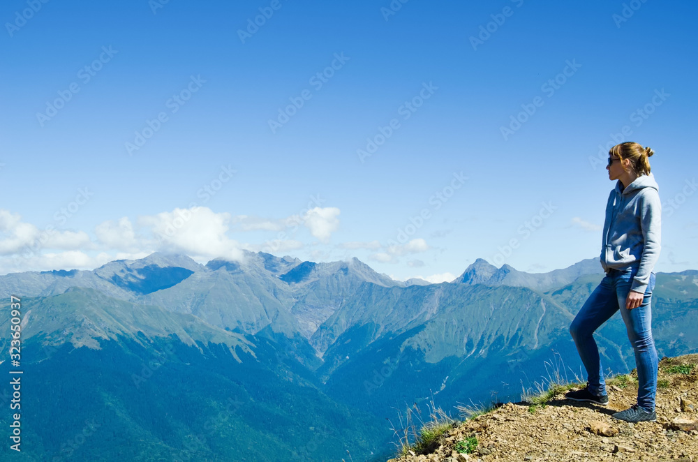 A woman in jeans and a sweatshirt stands on top of a mountain and looks at the mountains.