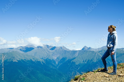 A woman in jeans and a sweatshirt stands on top of a mountain and looks at the mountains.