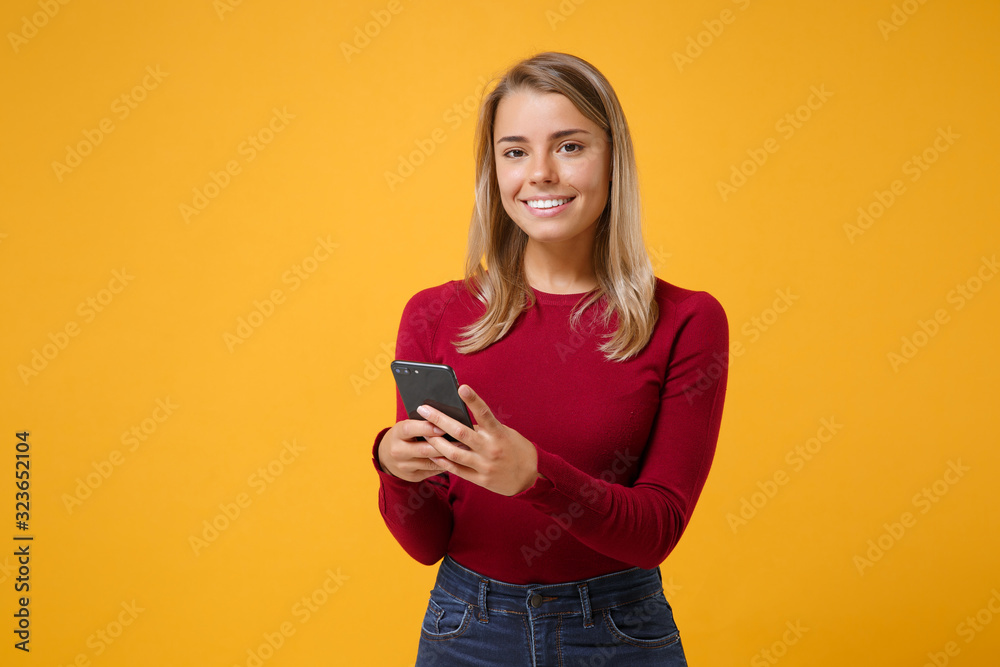 Smiling young blonde woman girl in casual clothes posing isolated on yellow orange wall background studio portrait. People lifestyle concept. Mock up copy space. Using mobile phone typing sms message.