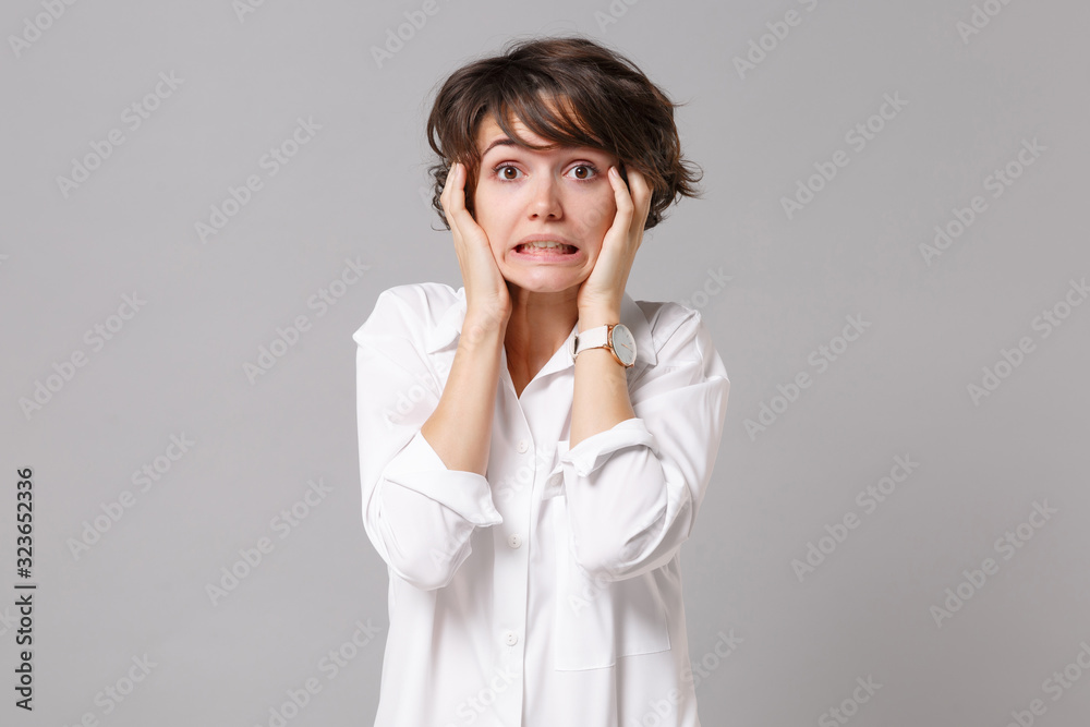 Confused puzzled young business woman in white shirt posing isolated on grey wall background studio portrait. Achievement career wealth business concept. Mock up copy space. Putting hands on cheeks. Confused puzzled young business woman in white shirt posing isolated on grey wall background studio portrait. Achievement career wealth business concept. Mock up copy space. Putting hands on cheeks.