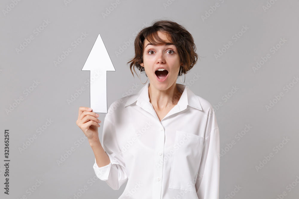 Shocked young business woman in white shirt posing isolated on grey wall background studio portrait. Achievement career wealth business concept. Mock up copy space. Hold up arrow, keeping mouth open. Shocked young business woman in white shirt posing isolated on grey wall background studio portrait. Achievement career wealth business concept. Mock up copy space. Hold up arrow, keeping mouth open.