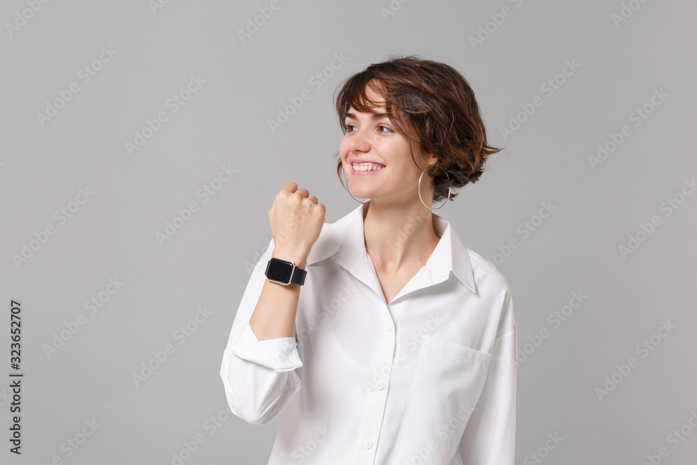 Smiling young business woman in white shirt posing isolated on grey background in studio. Achievement career wealth business concept. Mock up copy space. Wearing smart watch on hand, looking aside. Smiling young business woman in white shirt posing isolated on grey background in studio. Achievement career wealth business concept. Mock up copy space. Wearing smart watch on hand, looking aside.