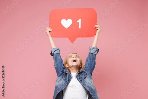 Cheerful little kid girl 12-13 years old in denim jacket isolated on pastel pink background in studio. Childhood lifestyle concept. Mock up copy space. Hold huge like sign from Instagram heart form. photo