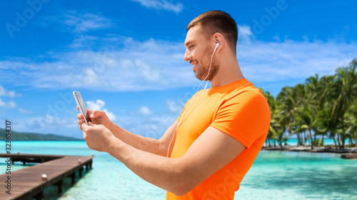 fitness, sport and people concept - smiling young man with smartphone and earphones listening to music at summer over wooden pier, palm trees and sea in french polynesia on background
