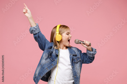Cute little kid girl 12-13 years old isolated on pastel pink background. Childhood lifestyle concept. Mock up copy space. Listen music with headphones sing song in microphone pointing index finger up.