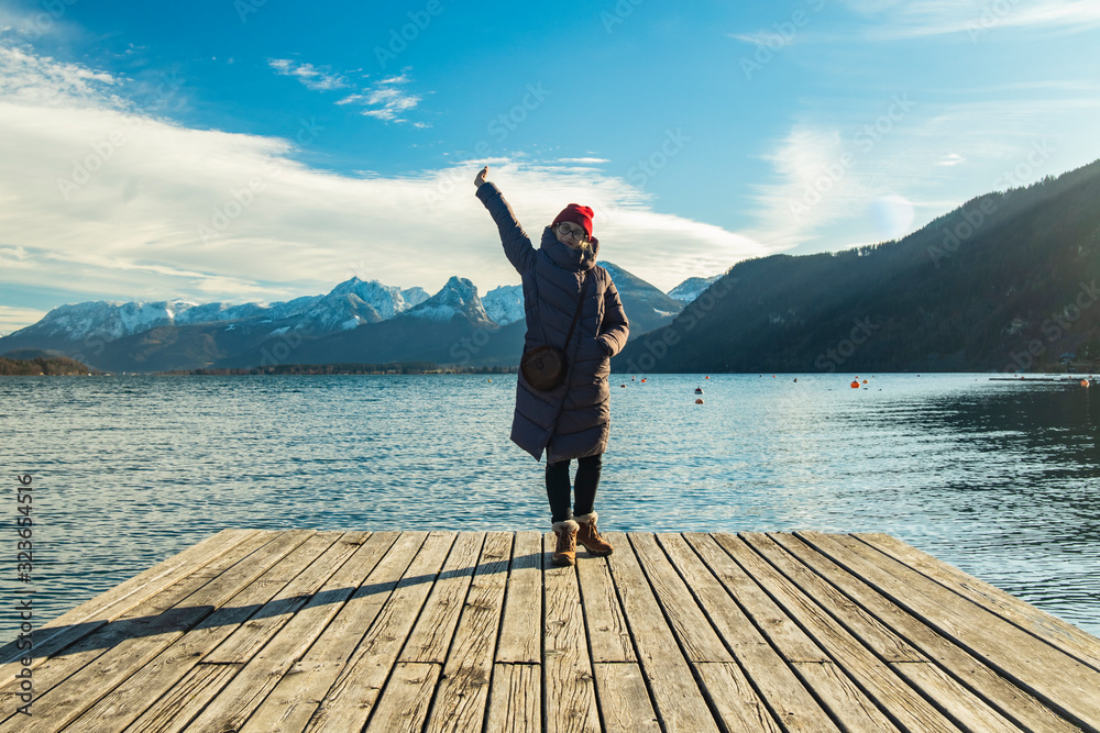 travel photography concept of woman posing portrait on wooden waterfront pier of lake waters and picturesque Alps mountains landscape background scenic view winter time