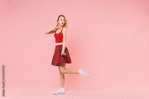Amazed young blonde woman girl in red sexy clothes isolated on pastel pink background. People lifestyle concept. Mock up copy space. Whispering secret behind her hand, sharing news, looking aside.