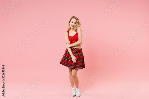 Smiling young blonde woman girl in red sexy clothes isolated on pastel pink wall background studio portrait. People emotions lifestyle concept. Mock up copy space. Looking down holding hands crossed.