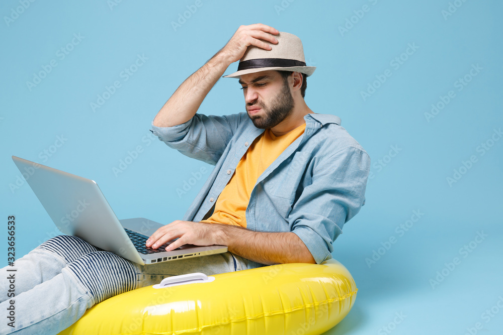 Puzzled traveler tourist man in yellow clothes isolated on blue background. Passenger traveling abroad on weekends. Air flight journey concept. Sit in inflatable ring, work on laptop, booking hotel.