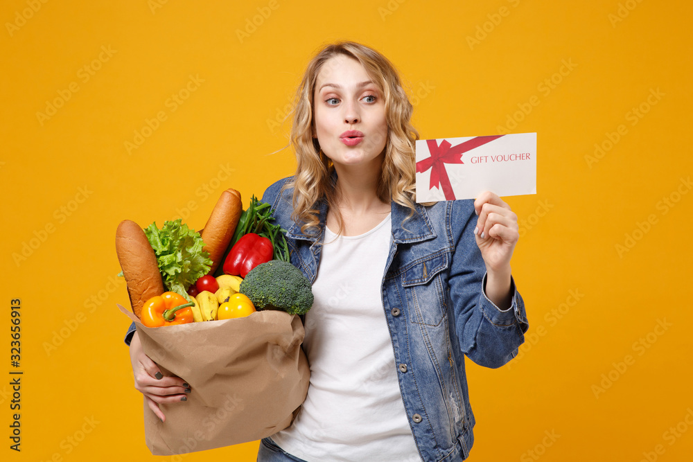 Amazed young woman in denim clothes isolated on orange background. Delivery service from shop or restaurant concept. Hold brown craft paper bag for takeaway mock up with food product gift certificate. Amazed young woman in denim clothes isolated on orange background. Delivery service from shop or restaurant concept. Hold brown craft paper bag for takeaway mock up with food product gift certificate.