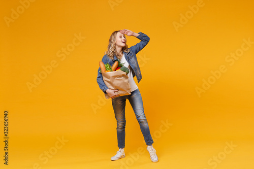 Funny girl in denim clothes isolated on orange background. Delivery service from shop or restaurant concept Hold brown craft paper bag for takeaway mock up with food product looking far away distance.