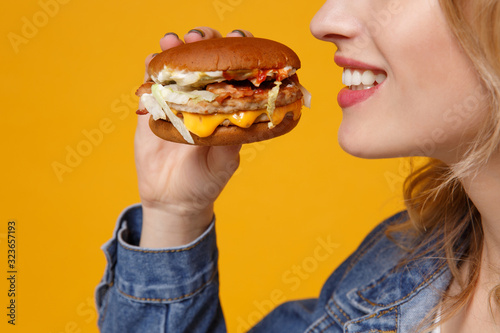 Cropped image side view of smiling young woman in denim clothes isolated on orange background studio portrait. Proper nutrition or American classic fast food concept. Mock up copy space. Hold burger.