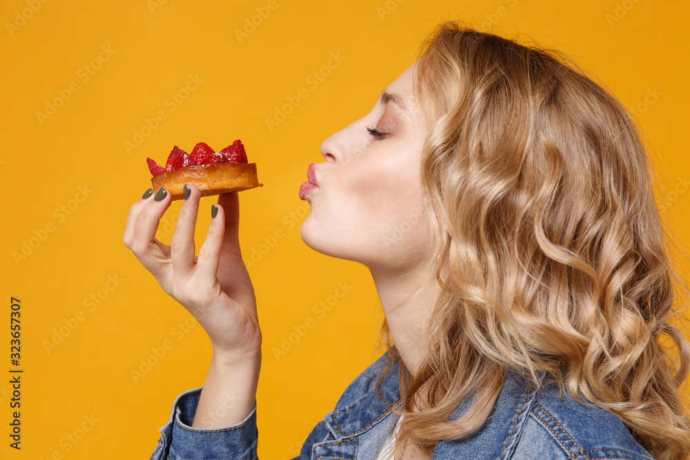 Side view of young woman in denim clothes isolated on yellow orange background in studio. Proper nutrition or sweets, dessert fast food, dieting concept. Mock up copy space. Eating strawberry cake. Side view of young woman in denim clothes isolated on yellow orange background in studio. Proper nutrition or sweets, dessert fast food, dieting concept. Mock up copy space. Eating strawberry cake.
