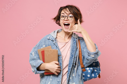 Excited young woman student in denim clothes glasses backpack isolated on pastel pink background. Education in high school university college concept. Hold books doing phone gesture says call me back. photo