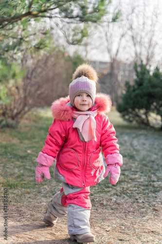 a three-year-old girl in a warm suit, hat and scarf walks along the first spring grass in the yard