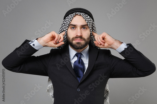 Young bearded arabian muslim businessman in keffiyeh kafiya ring igal agal classic black suit shirt isolated on gray background. Achievement career wealth business concept. Covering ears with fingers. photo