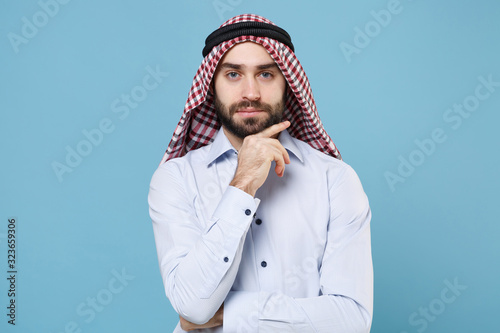 Handsome young arabian muslim man in keffiyeh kafiya ring igal agal casual clothes isolated on pastel blue background. People religious lifestyle concept. Mock up copy space. Put hand prop up on chin. photo