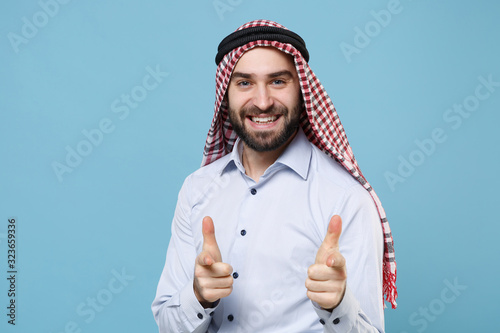 Smiling young bearded arabian muslim man in keffiyeh kafiya ring igal agal casual clothes isolated on pastel blue wall background. People religious lifestyle concept. Pointing index finger on camera. photo
