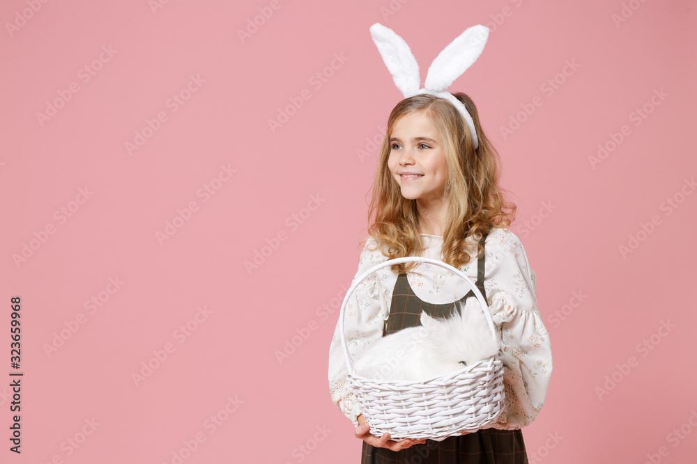 Little pretty blonde kid girl 11-12 years old in light spring dress hold wicker basket with cute fluffy white bunny rabbit isolated on pastel pink background. Childhood lifestyle Happy Easter concept. Little pretty blonde kid girl 11-12 years old in light spring dress hold wicker basket with cute fluffy white bunny rabbit isolated on pastel pink background. Childhood lifestyle Happy Easter concept.