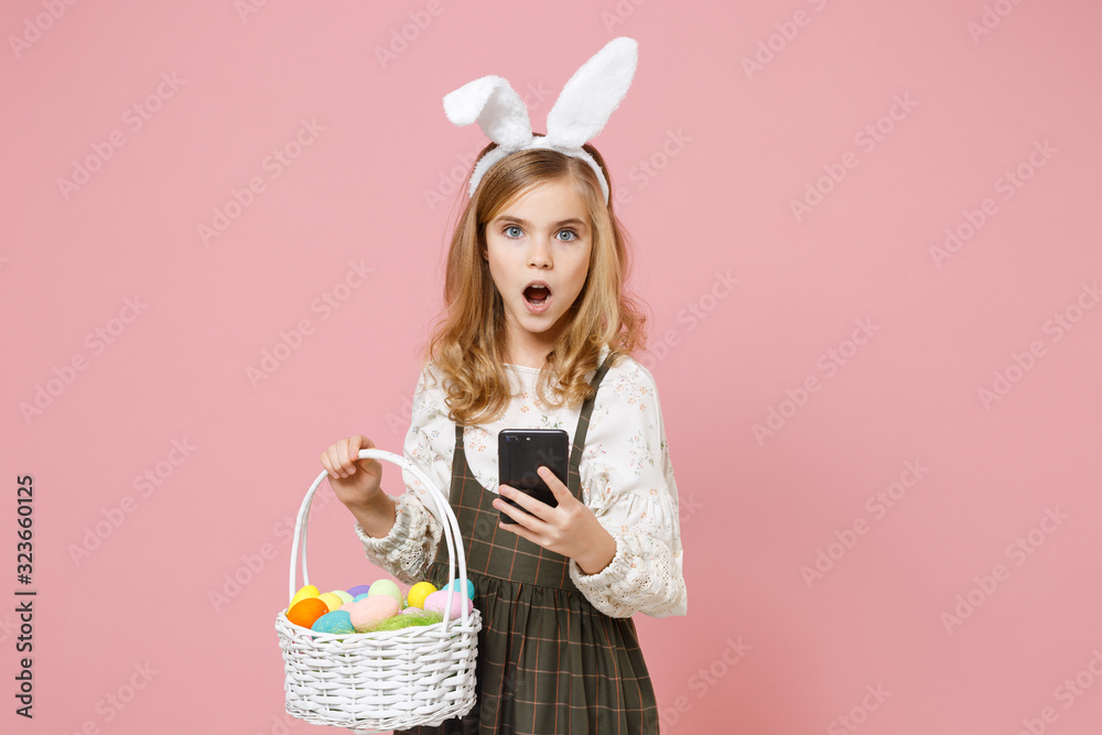 Little blonde kid girl 11-12 years old in spring dress bunny rabbit ears hold in hand cell phone wicker basket colorful eggs isolated on pastel pink background Childhood lifestyle Happy Easter concept Little blonde kid girl 11-12 years old in spring dress bunny rabbit ears hold in hand cell phone wicker basket colorful eggs isolated on pastel pink background Childhood lifestyle Happy Easter concept