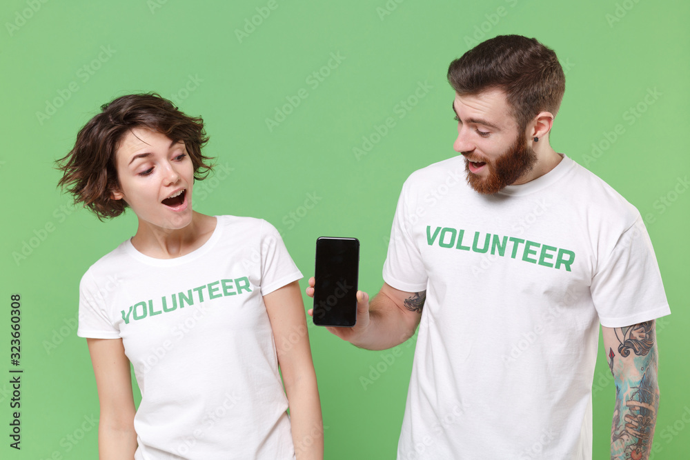 Shocked two young friends couple in white volunteer t-shirt isolated on green background. Voluntary free work assistance help charity grace teamwork concept. Hold mobile phone with blank empty screen. Shocked two young friends couple in white volunteer t-shirt isolated on green background. Voluntary free work assistance help charity grace teamwork concept. Hold mobile phone with blank empty screen.