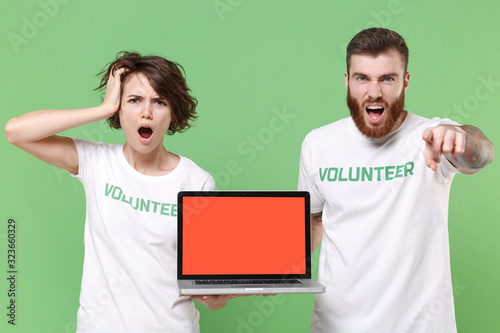 Preoccupied shocked two friends couple in volunteer t-shirt isolated on green background. Voluntary free work assistance help charity grace teamwork. Hold laptop pc computer with blank empty screen.