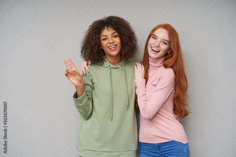 Joyful young lovely redhead woman with long hair embracing her cheerful curly dark skinned brunette friend and smiling widely while posing over grey background Joyful young lovely redhead woman with long hair embracing her cheerful curly dark skinned brunette friend and smiling widely while posing over grey background
