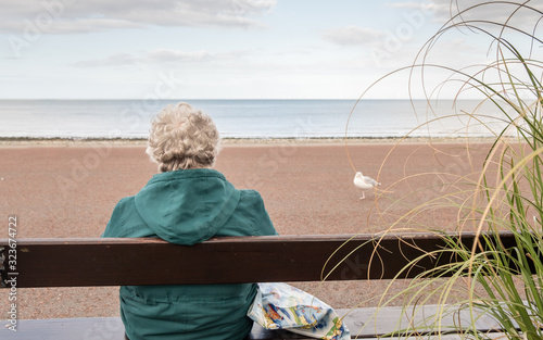 Old age - alone. Old woman with bag seen from behind sitting looking out to sea. Grandmother loneliness.