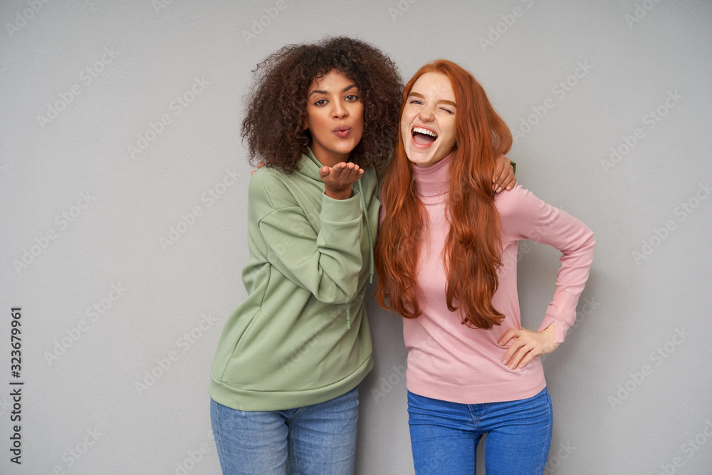 Joyful young lovely redhead woman with casual hairstyle winking happily at camera while embracing curly dark skinned brunette female blowing air kiss to camera, isolated over grey background Joyful young lovely redhead woman with casual hairstyle winking happily at camera while embracing curly dark skinned brunette female blowing air kiss to camera, isolated over grey background