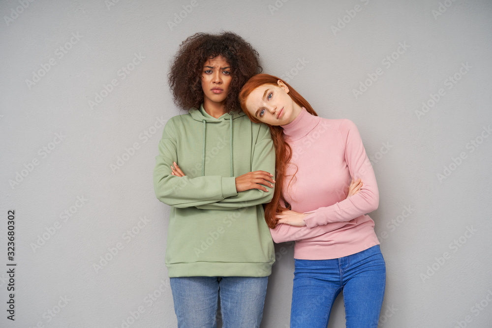 Puzzled young pretty brown haired dark skinned woman keeping her hands folded and looking sadly at camera while posing over grey background with her redhead calm friend Puzzled young pretty brown haired dark skinned woman keeping her hands folded and looking sadly at camera while posing over grey background with her redhead calm friend